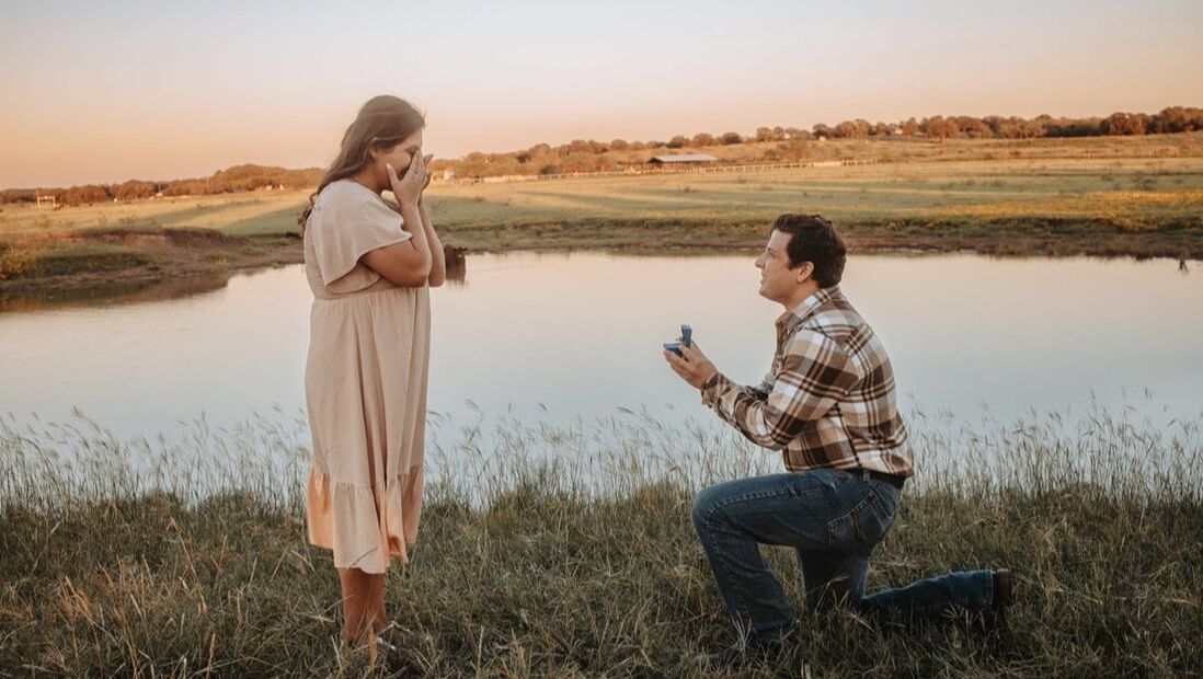 Jacob proposing to Jessica in a field next to a pond in the countryside.