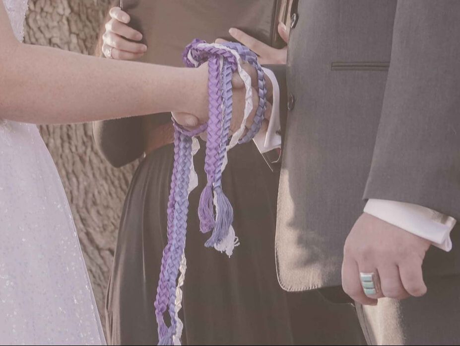 Bride and groom holding hands within purple, blue, and white handfasting ribbons
