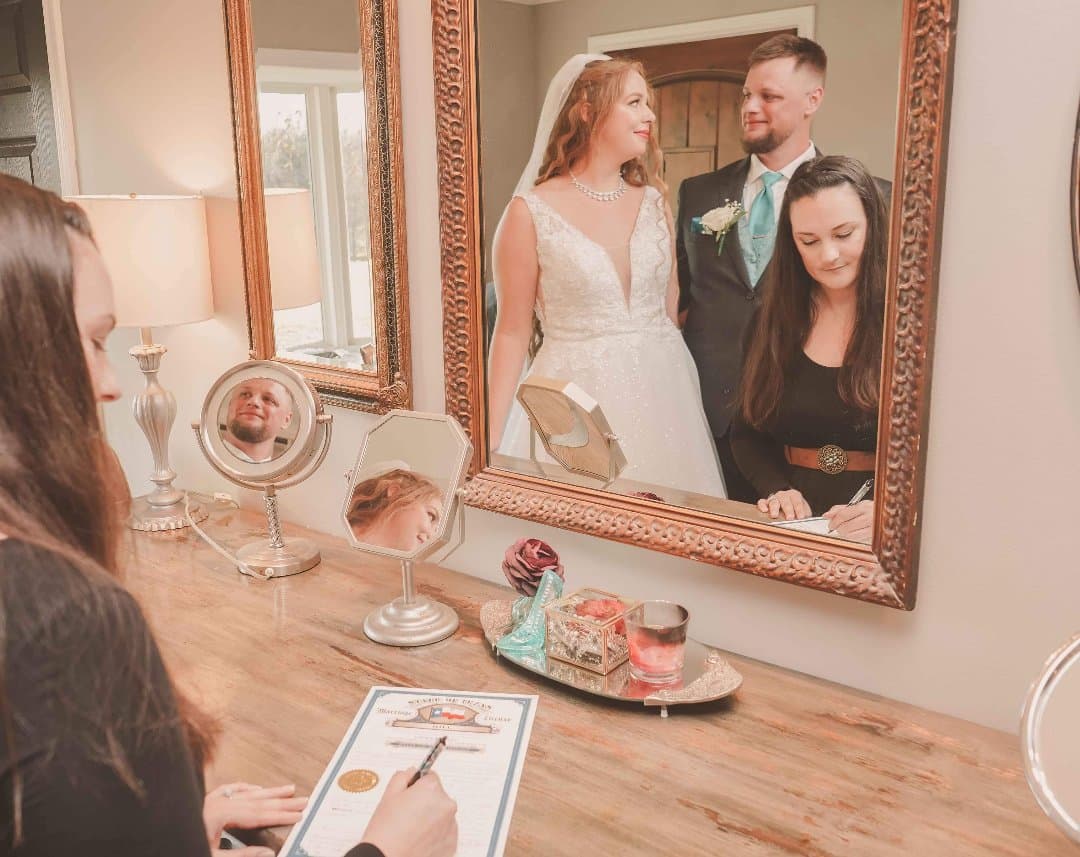 Reflection in a mirror of bride and groom looking at each other lovingly while the officiant signs the marriage license.